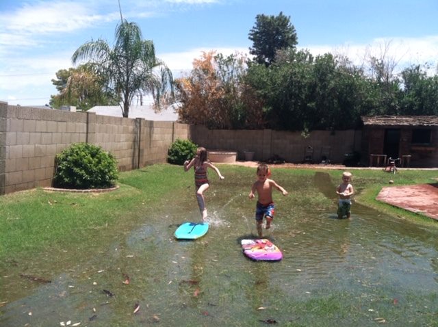 Image shows 3 children playing on short boogie board surrounded by shallow water